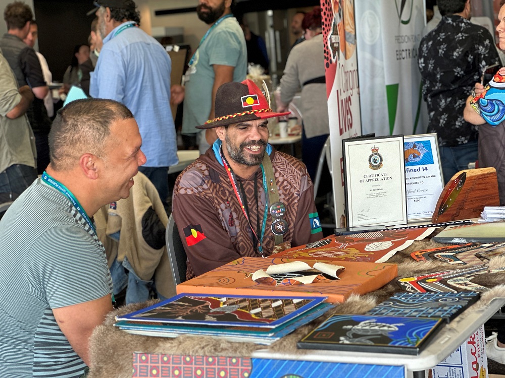 Two First Nations business owners sit at a booth smiling. The one closet to camera is facing away from the camera, while the one in the background is facing towards the camera, wearing a hat and top adorned with First Nations artwork and the Aboriginal flag.