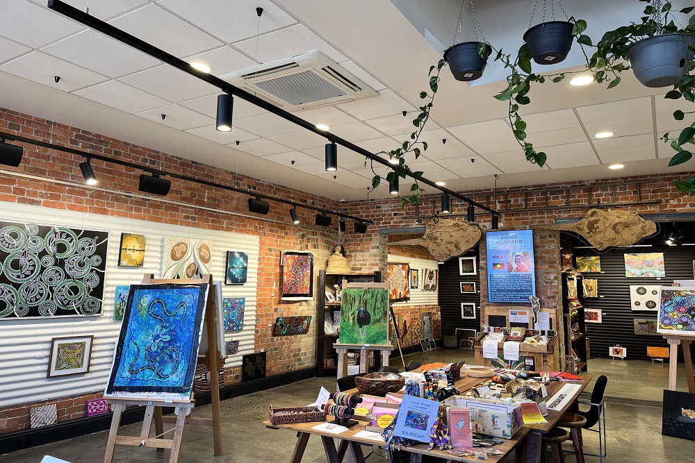 The interior of Perridak Arts gallery, featuring a range of Aboriginal artworks that are hung from the walls, mounted on stands or spread across tables. At the top right of the frame, hanging plants are visible. 