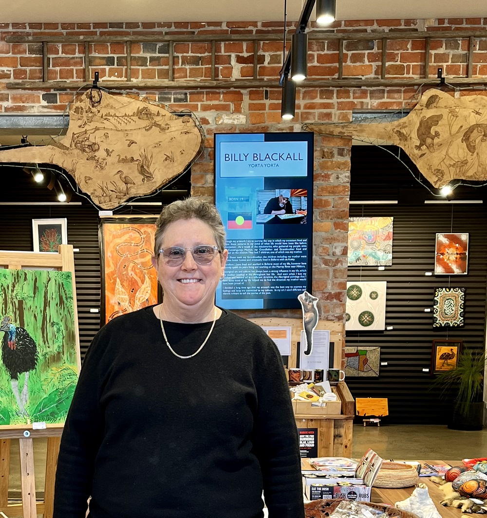 Perridak Arts gallery manager Sue Clark stands smiling at the camera, with a range of art displays visible behind her against a brick wall background.