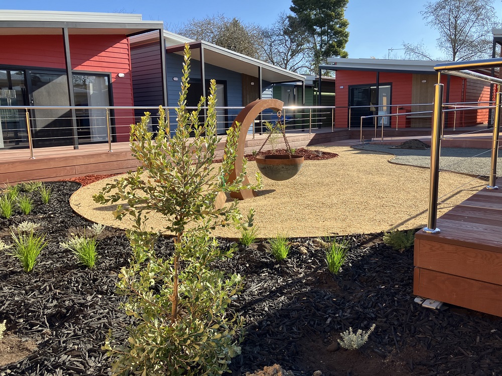 A courtyard containing small native bushes in brown tanbark can be seen in the foreground, with a wooden hanging plant stand visible in the centre of the frame. In the background are two red cabins with a blue cabin between them. A veranda with a silver handrail stretches around the courtyard, linking to the cabins.