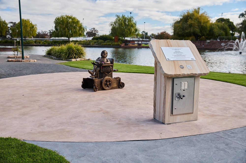 A bronze-coloured statue of Stella Young sits in the centre of a circular section of pathway, with a tall plaque visible in the foreground, and trees, lawn and a lake visible in the background.