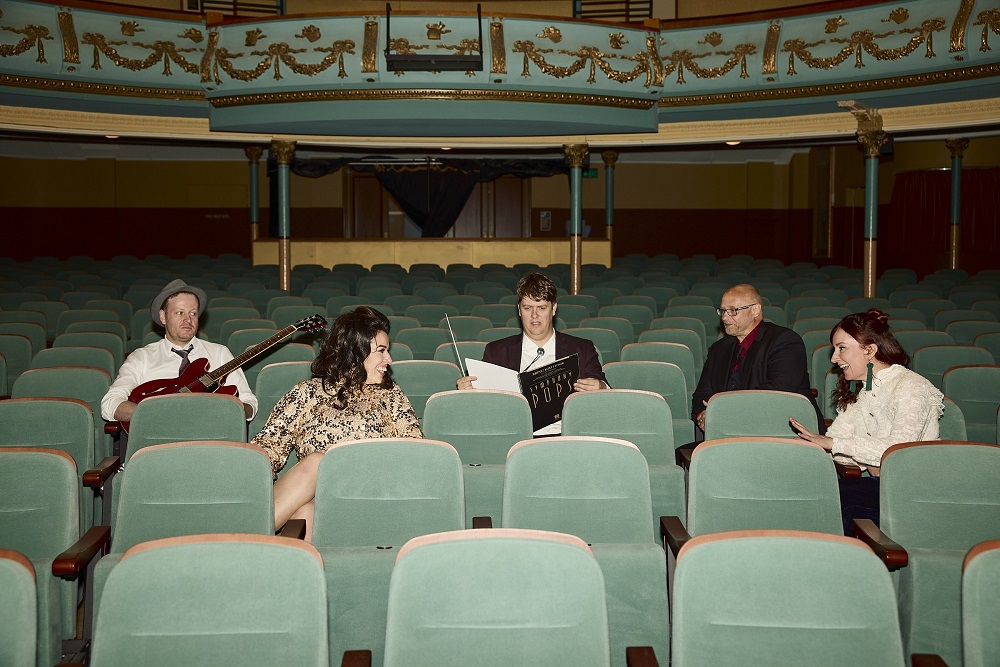 Five performers sitting in the green seats of Her Majesty's Theatre Ballarat's auditorium. The performers are smiling, holding their instruments and looking at each other. The balcony level of the theatre is visible at the top of the photo.