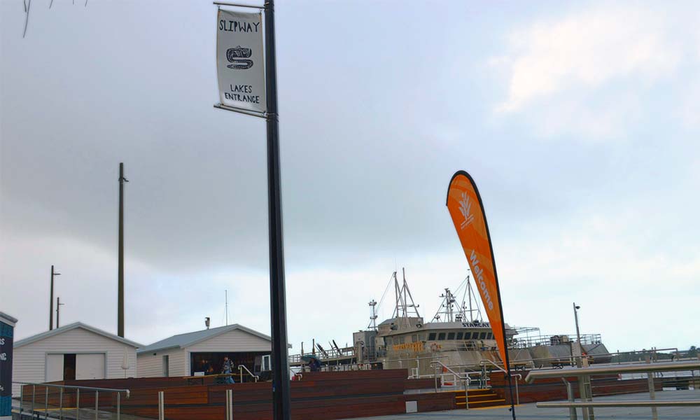 Two winch sheds are in the background next to a large fishing barge. There is a street pole with a banner that reads ‘slipway Lakes Entrance’.