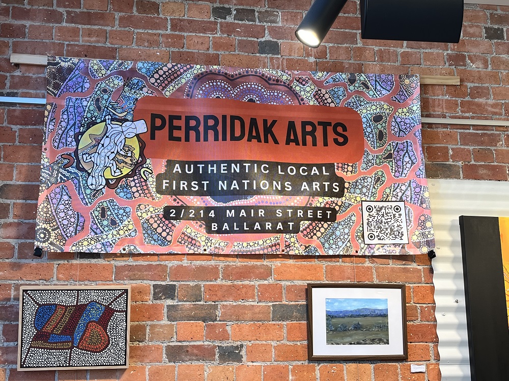 Perridak Arts sign visible in centre of frame, against a brick backdrop. In the foreground are visible pieces of Aboriginal art.