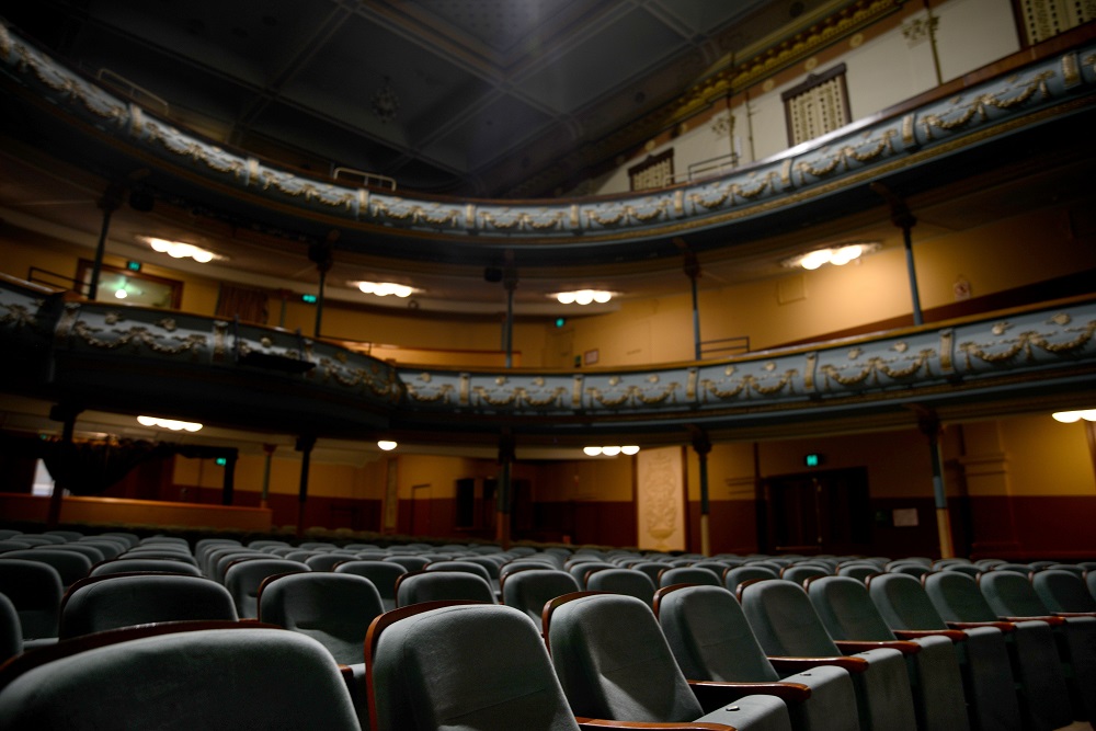 The auditorium of Her Majesty’s Theatre Ballarat, with empty blue seats folded up in the foreground. In the background the mezzanine and balcony are visible, with yellow theatre lighting illuminating the edges of the auditorium.