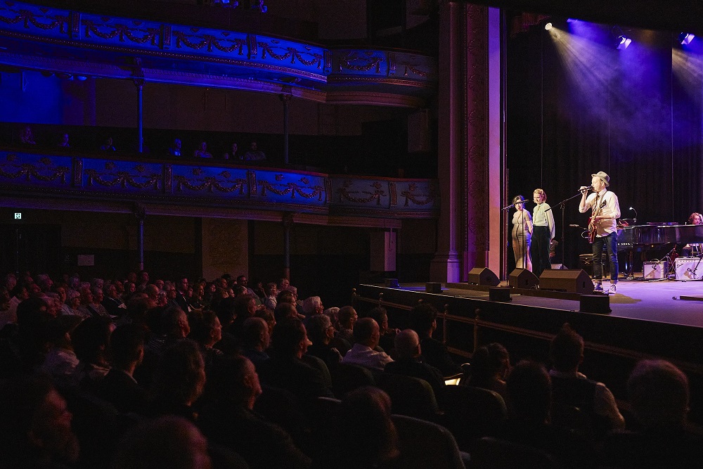 The 'Resurgence' performance at Her Maj. The audience and auditorium are bathed in purple lighting on the left of the image, while the performers are visible on stage to the right of the image.