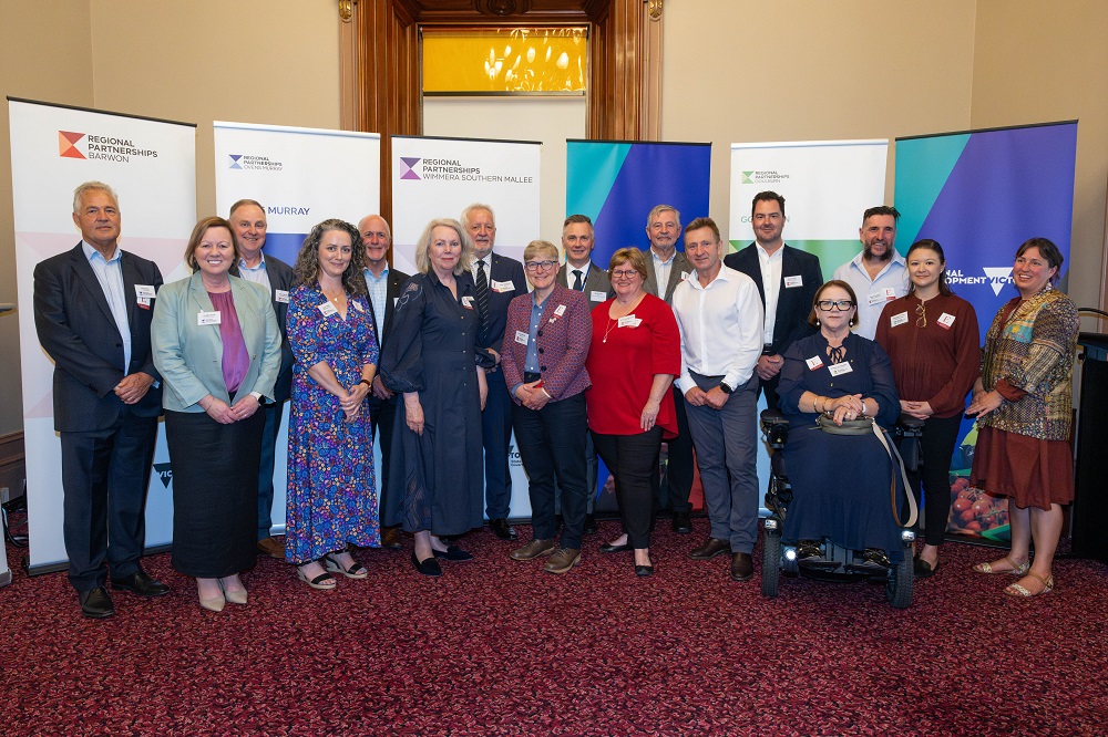 Men and women from the Regional Partnerships boards standing in front on branded banners, with red carpet visible in the foreground and cream coloured walls visible in the background.  