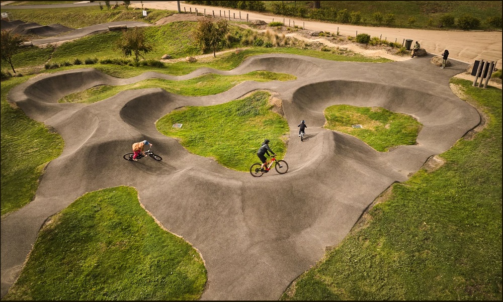 Three riders taking a corner on the Hammon Park Trailhead, an enclosed training area of the park. Photo taken from a bird's eye view, with the black jumps visible and moving across the image in a series of circular mini trails.