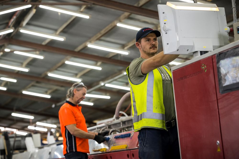 Two workers operating inside an industrial space. A worker wearing a green reflective vest is operating a computer in the right side of the image, while a worker in a red shirt operates a red machine in the background. White lights and a steel roof are visible at the top of the image.