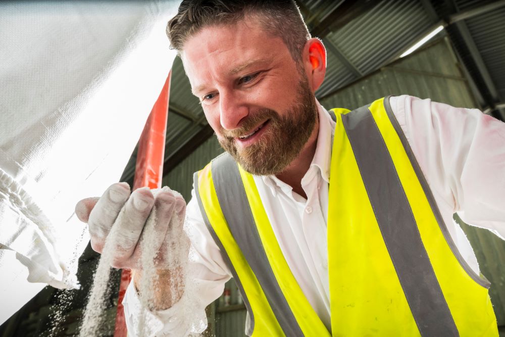 Jack Fitzgerald, wearing a white shirt and yellow reflective vest, smiles down at white dust-like material as it falls between his fingers.